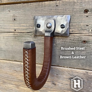 Hand-forged brushed steel gun hook wall mount wrapped in brown leather riveted to a hammered steel plate and mounted on a rustic wooden wall. Hutton Forge Brushed Steel & Brown Leather gun display hook.