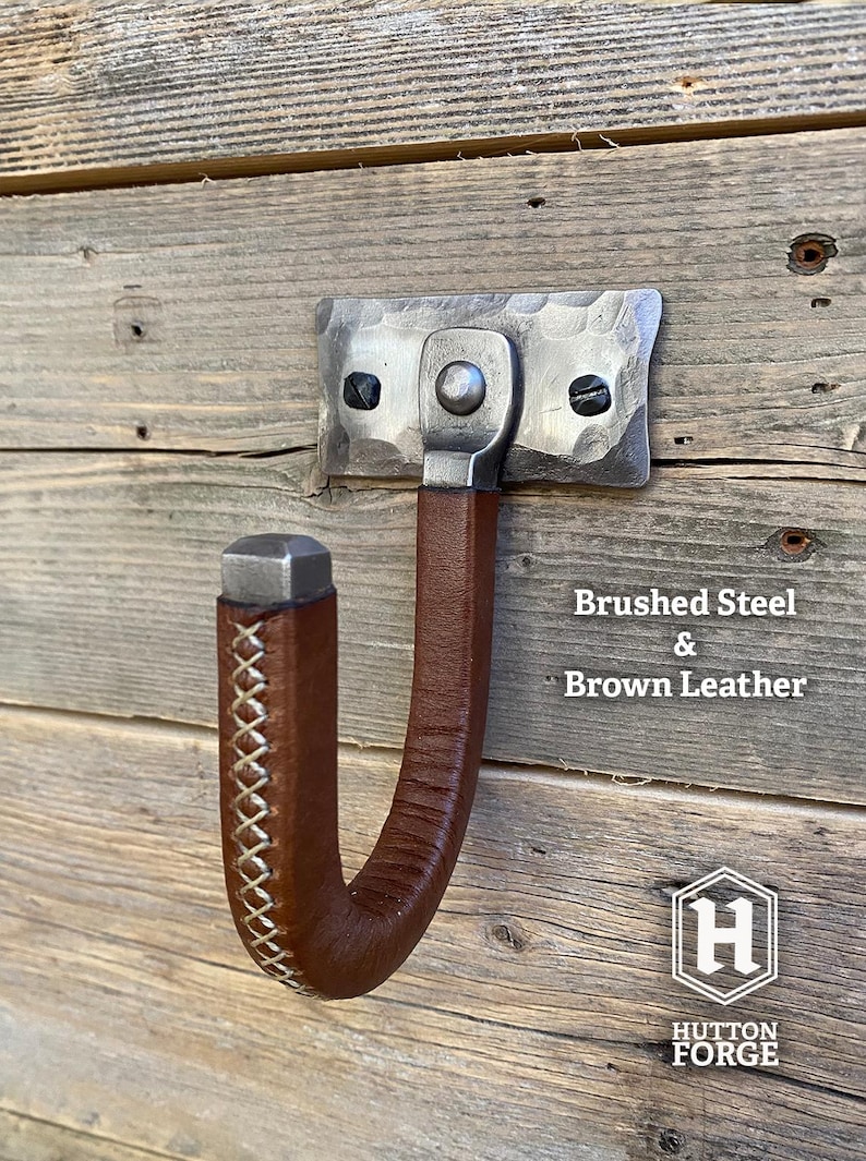 Hand-forged brushed steel gun hook wall mount wrapped in brown leather riveted to a hammered steel plate and mounted on a rustic wooden wall. Hutton Forge Brushed Steel & Brown Leather gun display hook.
