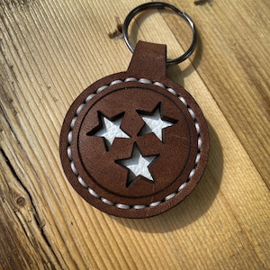 May include: A circular brown leather keychain featuring three star-shaped cutouts that reveal a white textured layer underneath. The leather piece is secured with a metal split ring and is finished with thick, light-colored stitching around the edge.