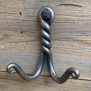May include: Hand-forged metal hook with a twisted central design and two curved hooks at the bottom. The hook is a dark gray color and appears to be made of iron. The hook is mounted on a wooden surface.