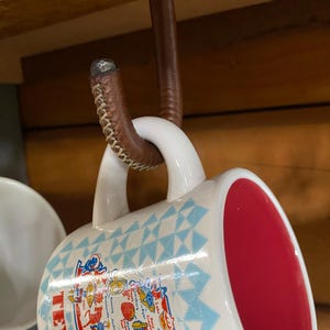May include: White ceramic mug with a red interior, featuring a map design with blue and red accents. The mug is suspended from a brown leather-wrapped hook attached to a wooden beam. The hook has a metal top.