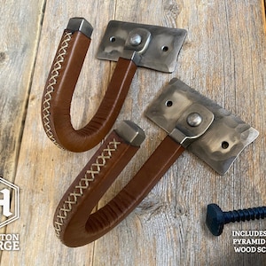 Hand-forged brushed steel J-hook wall mount wrapped in brown leather with cross-stitching, riveted to a hammered steel plate and mounted on a rustic wooden wall. Hutton Forge Brushed Steel & Brown Leather gun display hook.