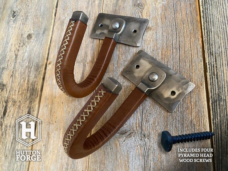 Hand-forged brushed steel J-hook wall mount wrapped in brown leather with cross-stitching, riveted to a hammered steel plate and mounted on a rustic wooden wall. Hutton Forge Brushed Steel & Brown Leather gun display hook.