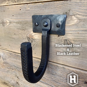 Hand-forged blackened steel gun hook wall mount wrapped in black leather, riveted to a hammered steel plate and mounted on a rustic wooden wall. Hutton Forge Blackened Steel & Black Leather gun display hook.