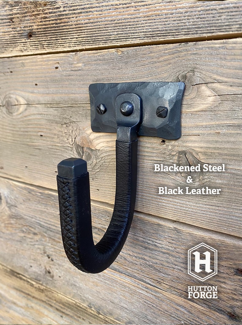 Hand-forged blackened steel gun hook wall mount wrapped in black leather, riveted to a hammered steel plate and mounted on a rustic wooden wall. Hutton Forge Blackened Steel & Black Leather gun display hook.