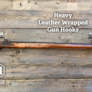 A leather-wrapped gun hook mounted on a rustic wooden plank wall, with a rifle resting horizontally across it for demonstration. Brushed Steel & Brown Leather gun display hook made by Hutton Forge.