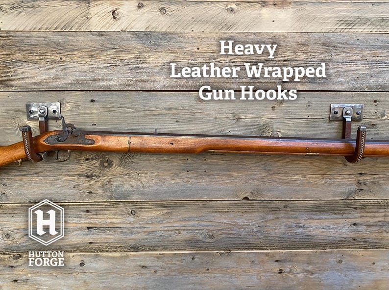 A leather-wrapped gun hook mounted on a rustic wooden plank wall, with a rifle resting horizontally across it for demonstration. Brushed Steel & Brown Leather gun display hook made by Hutton Forge.