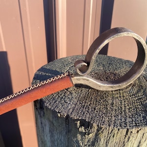 Puede incluir: Mango de metal forjado a mano con empuñadura de cuero marrón. El mango presenta un diseño circular con un remolino decorativo. La empuñadura de cuero está cosida con hilo blanco. El mango descansa sobre un poste de madera desgastado.