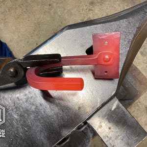 A blacksmith's forge with a glowing red metal gun hook being shaped on an anvil. The hook is held by tongs, and the Hutton Forge logo is visible in the lower left corner.