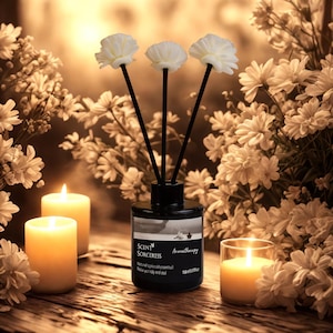 May include: A black aromatherapy diffuser with three white flower-shaped reeds. The diffuser is labeled "Scent Sorceress" and sits on a wooden surface with lit candles and white flowers in the background. The image has a warm, inviting aesthetic.