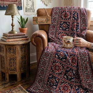 May include: A throw blanket featuring a repeating grid pattern of dark blue and deep red floral motifs. The blanket is draped over a brown leather armchair. A ceramic mug with a camel illustration and the text Sindh is held in the foreground.