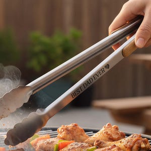 Op de afbeelding: Roestvrijstalen barbecuetang met houten handvatten, gegraveerd met "BEST DAD EVER". De tang houdt gegrilde kip- en groentespiesjes vast op een houtskoolgrill. Rook stijgt op van de grill, met een houten picknicktafel op de achtergrond.
