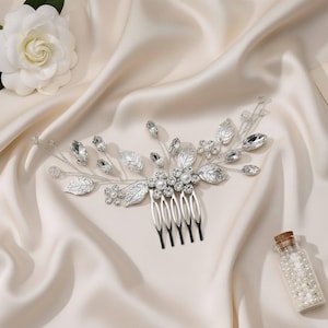 Puede incluir: Un peine de pelo plateado para novia, adornado con cristales transparentes, perlas de imitación y detalles de hojas. El peine se muestra sobre satén crema, con una flor blanca y un pequeño frasco de perlas. Accesorio de boda clásico.