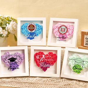 May include: Five framed heart-shaped floral arrangements in blue, pink, purple, red, and green. Each frame features a university emblem, a name, and the text "Class of 2026" or "Congratulations". A vase of white flowers is in the background.