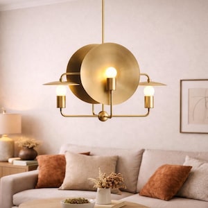 Könnte beinhalten: Eine moderne, goldfarbene Hängeleuchte mit drei Lichtern. Die Leuchte hat ein kreisförmiges Design mit drei Glühbirnen. Die Leuchte hängt über einem beigen Sofa mit orangefarbenen Kissen. Der Raum hat eine neutrale Farbpalette.