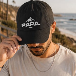 Könnte beinhalten: Schwarze Baseballkappe mit dem weißen Wort "PAPA" und einem Bergmotiv. Das Jahr "2026" ist unter dem Wort "PAPA" aufgedruckt. Die Kappe wird von einer Person getragen, die ein helles T-Shirt trägt.