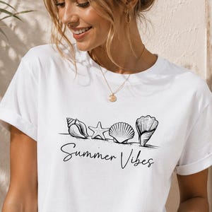 Könnte beinhalten: Ein weißes kurzärmeliges T-Shirt mit einem schwarzen Grafikdruck von vier verschiedenen Muscheln, die in einer Reihe über dem handschriftlichen Text Summer Vibes angeordnet sind. Das T-Shirt hat einen Rundhalsausschnitt, umgekrempelte Ärmel und eine hellblaue Jeansshorts.
