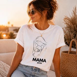 May include: White t-shirt with a black line drawing of a mother holding a baby. The word "MAMA" is printed below the image, with "2026" underneath. The shirt has rolled-up sleeves and is worn by a person.