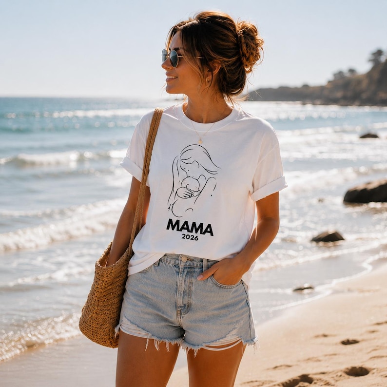 May include: White t-shirt with a black line drawing of a mother and child, with the word "MAMA" and the year "2026" printed below. The shirt is worn with denim shorts and a straw tote bag.