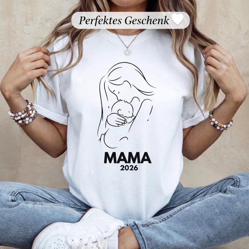 May include: White t-shirt featuring a black line drawing of a mother holding a baby, with the word "MAMA" and the year "2026" printed below. The text "Perfektes Geschenk" is above the drawing.