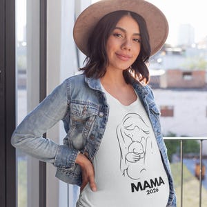 May include: A white t-shirt with a black line drawing of a mother holding a baby. The word "MAMA" and the year "2026" are printed in black below the illustration. The person is wearing a denim jacket and a brown hat.