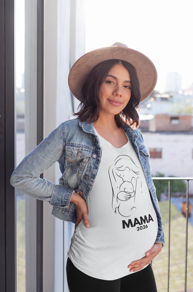May include: A white t-shirt with a black line drawing of a mother holding a baby. The word "MAMA" and the year "2026" are printed in black below the illustration. The person is wearing a denim jacket and a brown hat.