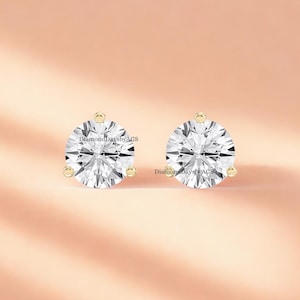 Puede incluir: Un par de pendientes de diamantes redondos con engastes de oro. Los diamantes claros y brillantes se destacan sobre un fondo rosa suave. Los pendientes se presentan en un estudio, mostrando su brillo.