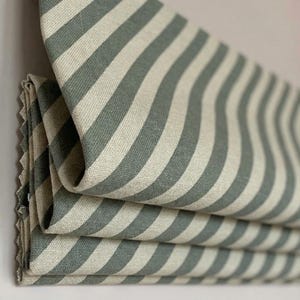May include: Close-up of folded fabric featuring broad, alternating stripes in sage green and cream. The material has a textured finish and is arranged in layered folds, highlighting the striped pattern. The edges are neatly finished.