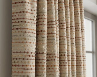 May include: Beige and brown textured curtains with horizontal rows of small, raised dots. The fabric is a neutral color, with varying shades of brown and beige. The curtains are hanging in vertical folds, creating a soft, flowing appearance.