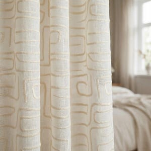 Puede incluir: Paneles de cortina color crema con un patrón geométrico repetitivo en un tono beige más claro. La tela tiene una apariencia texturizada y el diseño está bordado. Las cortinas cuelgan frente a una ventana, con una cama visible en el fondo.