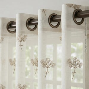 May include: Sheer white curtains with embroidered floral designs. The curtains have bronze grommets and are hung on a bronze rod. The text "NATURE'S WEAVE - EMBROIDERED SHEER" is printed on the side of the curtain.