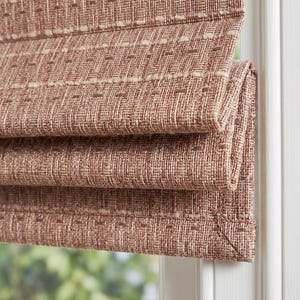 May include: A roman shade window covering featuring a textured, woven fabric in a warm terracotta brown hue. The material displays a subtle grid pattern with horizontal rows of lighter beige stitching, creating a layered, folded appearance.