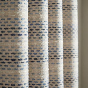 Puede incluir: Primer plano de cortinas de tela texturizada en color beige neutro con un patrón de pequeños puntos azules espaciados uniformemente. La tela es gruesa y parece estar tejida, con una textura ligeramente rugosa. Las cortinas están plegadas en pliegues verticales.