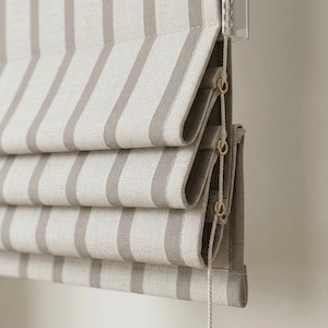 Puede incluir: Primer plano de una cortina romana con un patrón de rayas. La tela es de color neutro con rayas grises verticales. La cortina está parcialmente recogida, revelando la construcción plegada y el mecanismo de cordón.