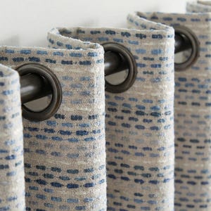 May include: Close-up of textured curtains with a neutral beige background and blue dot pattern. The curtains have dark bronze grommets for hanging. The fabric has a woven appearance, adding a touch of texture and visual interest to the design.