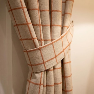 May include: Beige and orange plaid curtains with a matching tieback. The curtains are made of a textured fabric and feature a grid pattern in orange. The tieback is wrapped around the curtains to hold them in place.