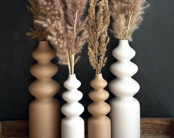 May include: Four decorative vases in varying shades of beige and white, each with a unique stacked design. The vases hold tall, feathery, dried pampas grass. The arrangement is set on a wooden tray against a dark background.