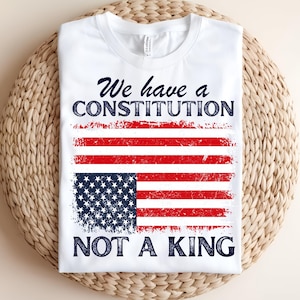 Op de afbeelding: Wit T-shirt met een verweerd ontwerp van de Amerikaanse vlag in rood, wit en blauw. De tekst "We have a Constitution, Not a King" is gedrukt in marineblauw. Een patriottisch kledingstuk.