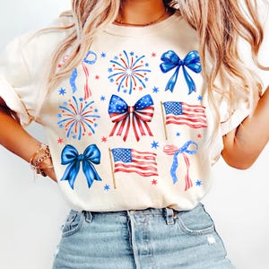 May include: Cream-colored t-shirt featuring patriotic designs. The shirt displays illustrations of blue bows, fireworks, American flags, and stars in red, white, and blue. The design evokes a festive, Independence Day theme.
