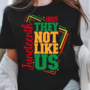 May include: Black t-shirt with the text "Juneteenth" in red script on the left side. The shirt features the words "1865 THEY NOT LIKE US" in green, orange, and black, with yellow and red accents. Stars are scattered around the text.