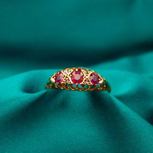 May include: A gold ring with three red gemstones set in a decorative filigree design. The ring is displayed on a teal-colored satin fabric, creating a luxurious and elegant presentation. This ring is a piece of jewelry.
