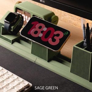May include: Sage green desk tidy with a built-in phone stand, watch holder, and pen holder. The phone displays the time 18:03. A keyboard and monitor are in the background. The organiser is made of a ribbed material.