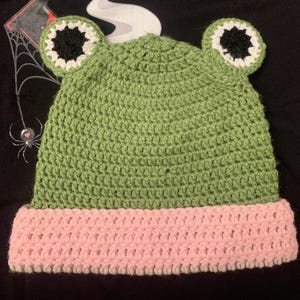 May include: A handmade crochet frog hat in green and pink. The hat features large, round eyes with white and black detailing. The brim is a soft pink, and the body of the hat is a textured green. The hat is designed to be worn.