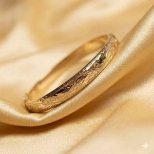 May include: A gold-colored bangle bracelet with intricate floral and leaf patterns. The bracelet is resting on a gold satin fabric, creating a luxurious and elegant display. The bangle is a classic piece of jewelry.