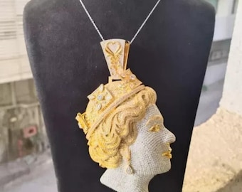 May include: A gold and silver pendant necklace featuring a profile of a person's face. The pendant has a detailed design with gold hair and a crown, and is encrusted with small, sparkling stones. The necklace is displayed on a black mannequin.