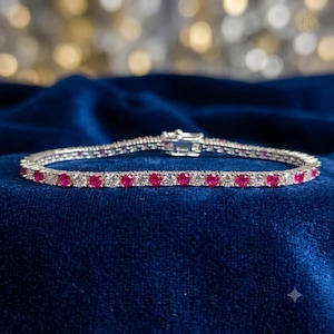 May include: A tennis bracelet featuring a repeating pattern of round-cut red rubies and clear white diamonds set in a silver-toned metal band. The bracelet includes a secure box clasp mechanism at the center, showcasing a continuous line of stones.