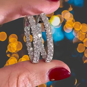 May include: A pair of silver hoop earrings encrusted with numerous small, sparkling diamonds. The earrings are held up against a blurred background of yellow and blue lights. The diamonds reflect light, creating a brilliant display.