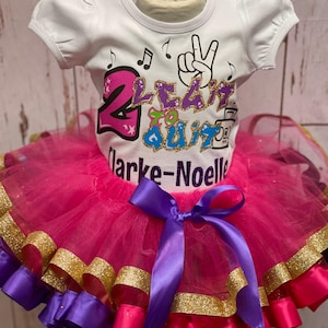 May include: A white short-sleeved top with the text "2 Lean to Quit" and musical notes, paired with a pink tutu skirt. The tutu has gold trim and a purple bow.