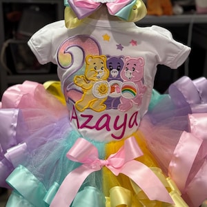 May include: A white short-sleeved bodysuit featuring an embroidered Care Bears graphic, the number two, and the name Azaya in pink script. The outfit includes a multi-colored pastel tulle tutu with a pink satin bow and a matching hair bow accessory.