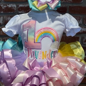 May include: A white short-sleeved shirt with a rainbow and the number 1 embroidered on the front. The shirt is paired with a multi-colored tutu skirt and a matching bow headband. The word "Lavender" is embroidered on the shirt.